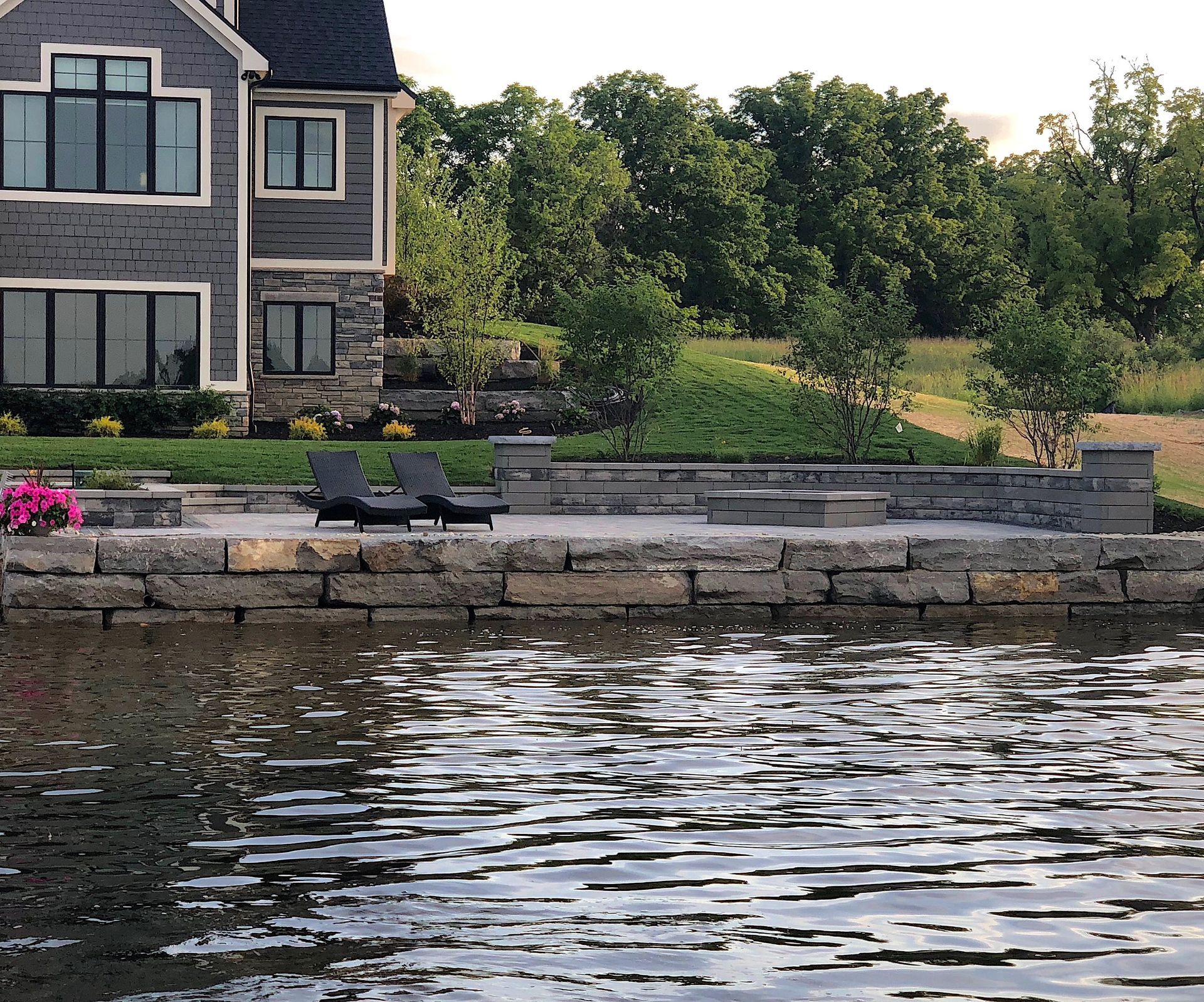 Lakeside house with stone patio and two lounge chairs, overlooking water; trees in the background.