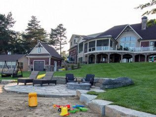 Lakeside home with stone patio, fire pit, and lawn; beach toys in the foreground, two-story house in the background.