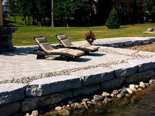Two lounge chairs on a stone patio overlooking a lake, with stone retaining walls and grass.