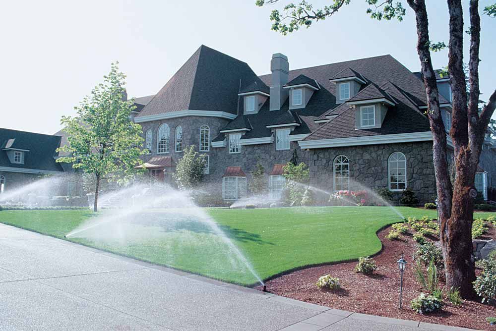 Sprinklers watering a green lawn in front of a large stone house.