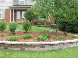 Front yard with a brick retaining wall, grass, plants, and a tree in front of a house.