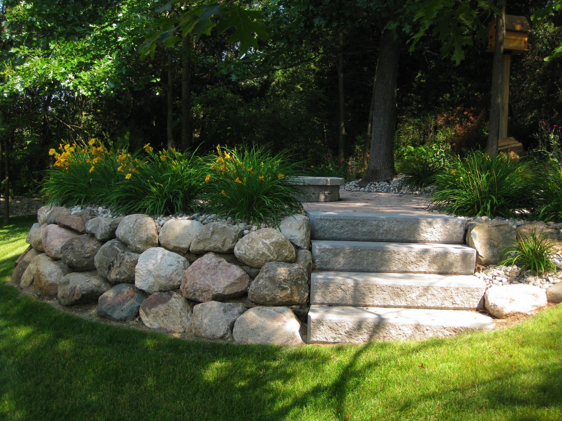 Stone retaining wall with steps, flowers, and path leading to a wooded area.