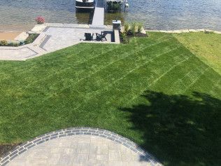 Lush green lawn with diagonal mowing pattern leading to a paved patio, dock, and lake.