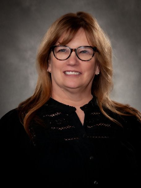 Woman with glasses smiles; wearing a black shirt, posing for a portrait against a gray background.