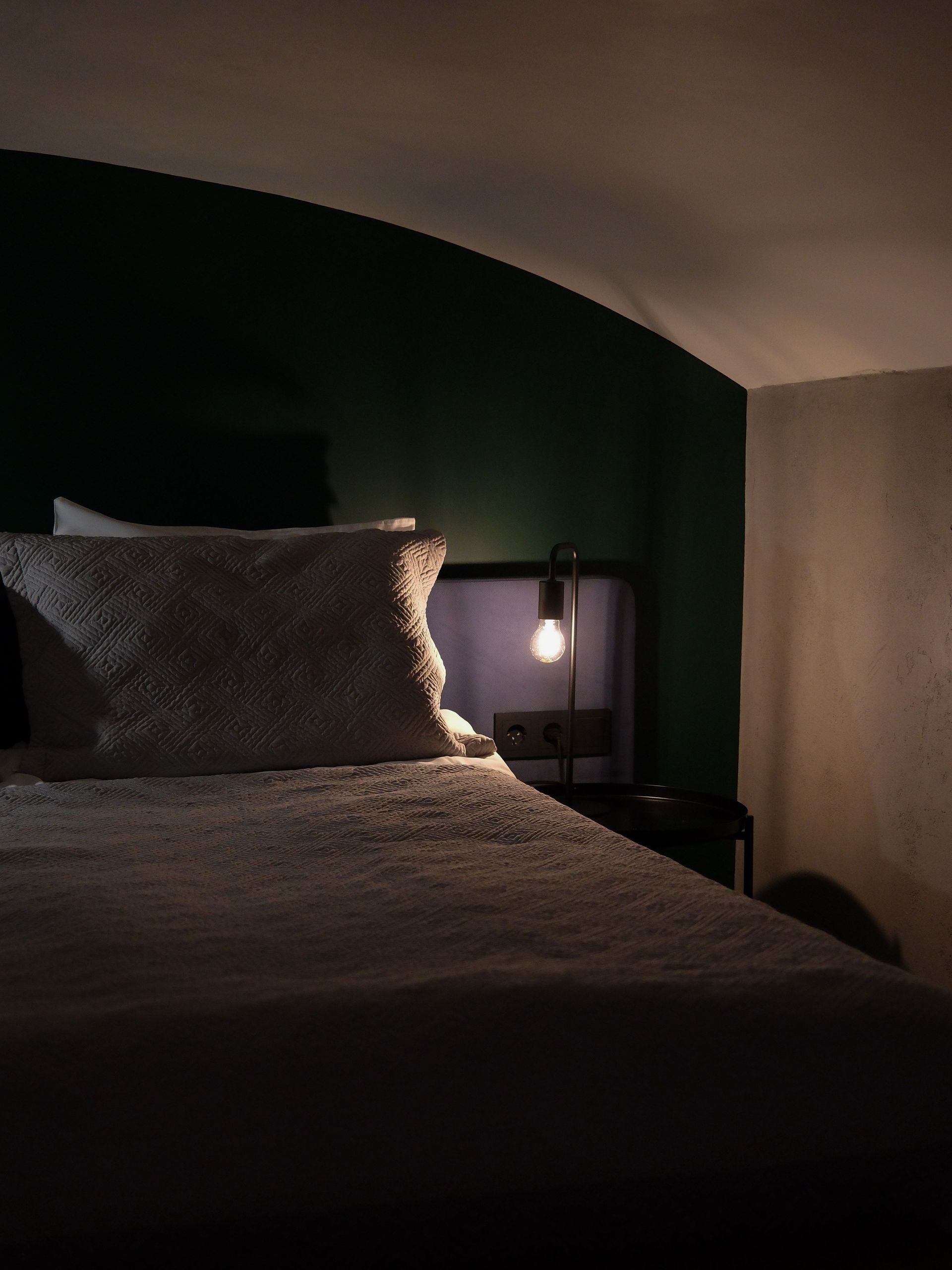 Dim bedroom with a bed, dark green wall, and a lit bedside lamp.
