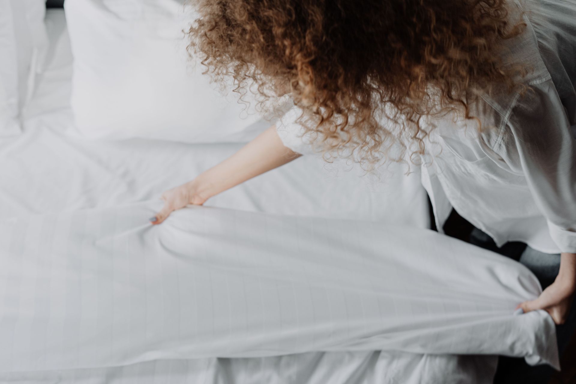 Person smoothing a white sheet on a bed in a bright bedroom