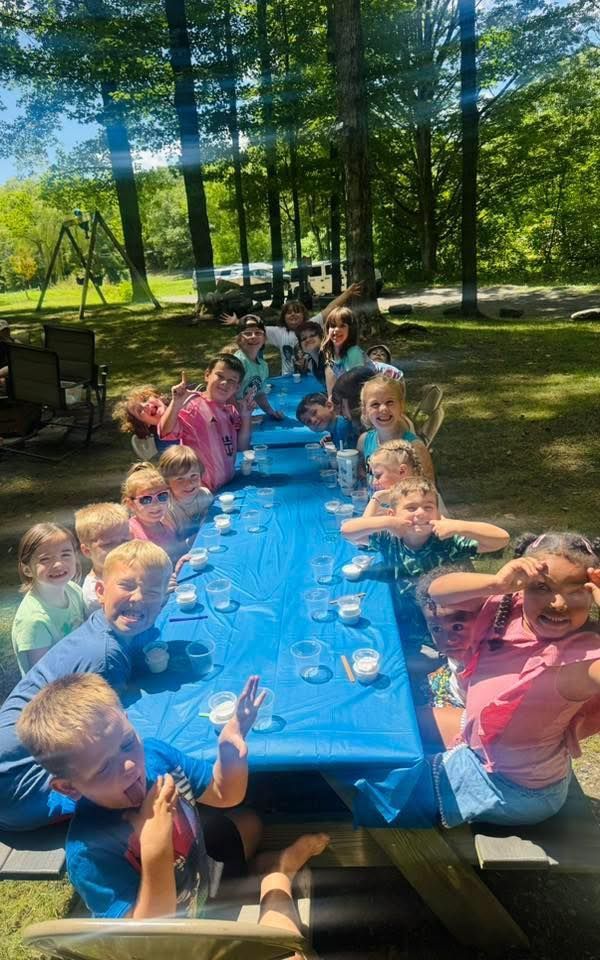 Children at a picnic table outside, smiling and making faces. Blue tablecloth, sunny day.