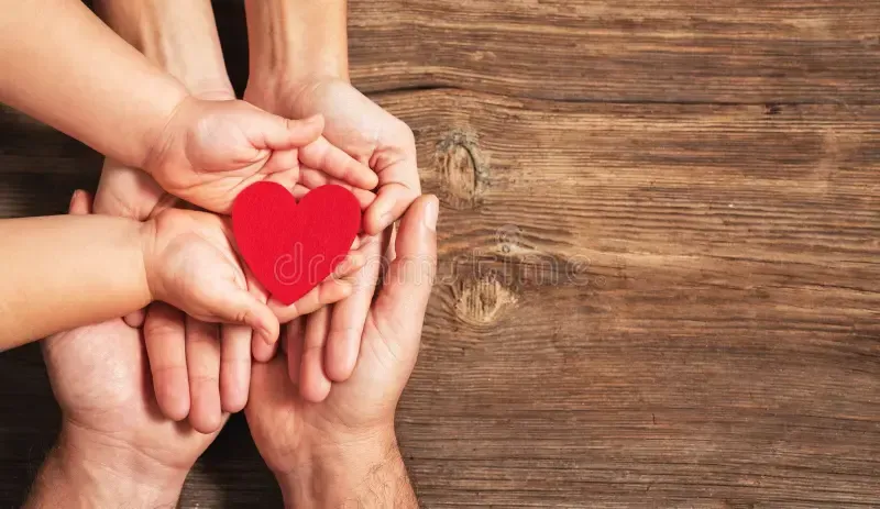 Hands of adults and a child holding a red heart on a wooden surface, symbolizing care.