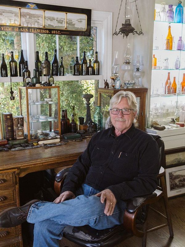 Man seated in a cozy room with a wine display and bottles on shelves behind him