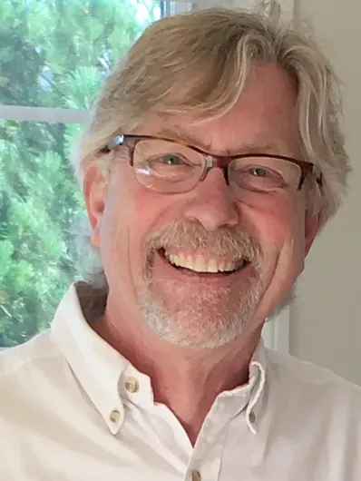Smiling man with glasses and gray hair, wearing a white shirt, in front of a window with greenery.