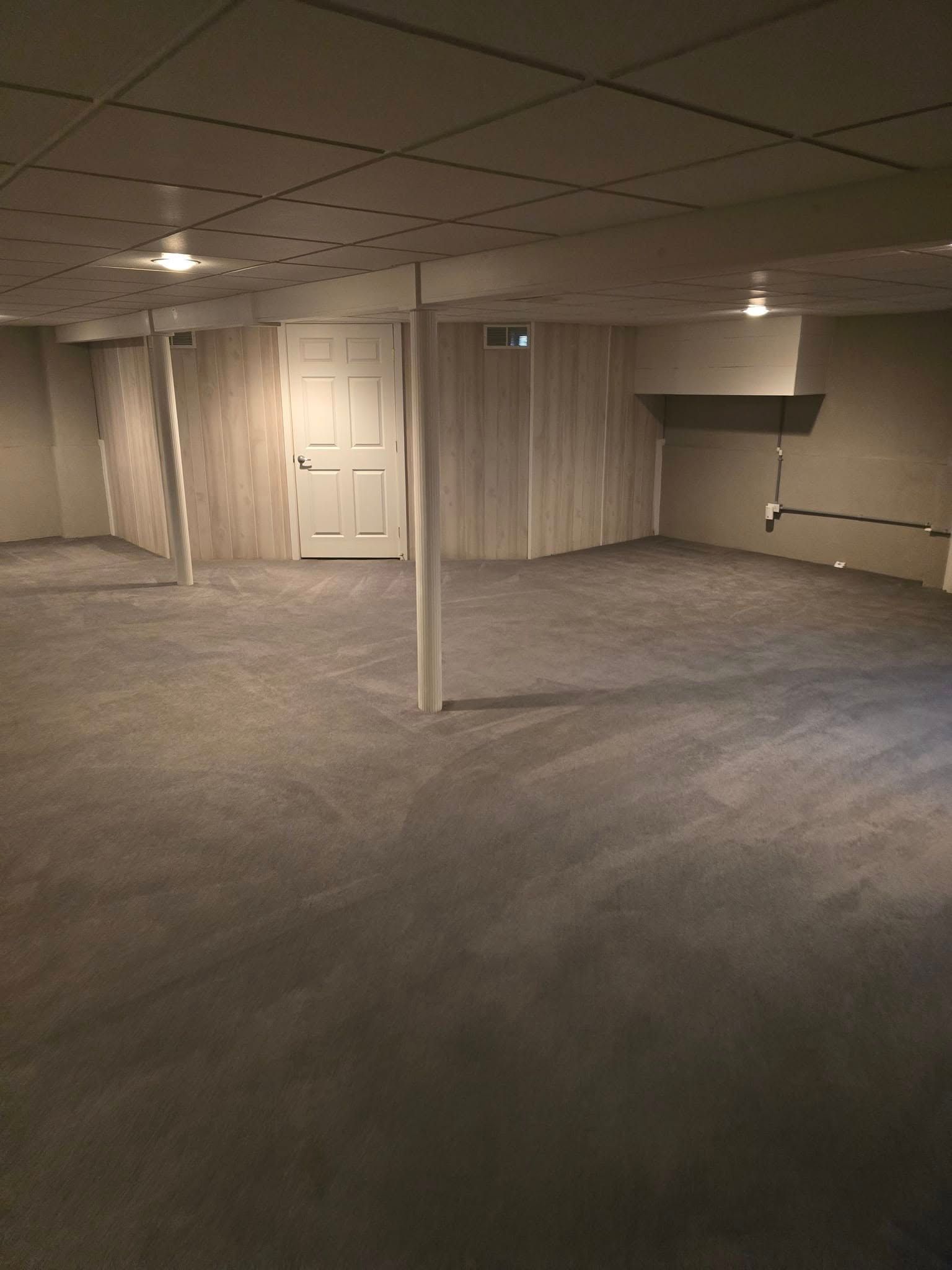 Empty carpeted basement room with beige paneled walls, ceiling tiles, and a support column