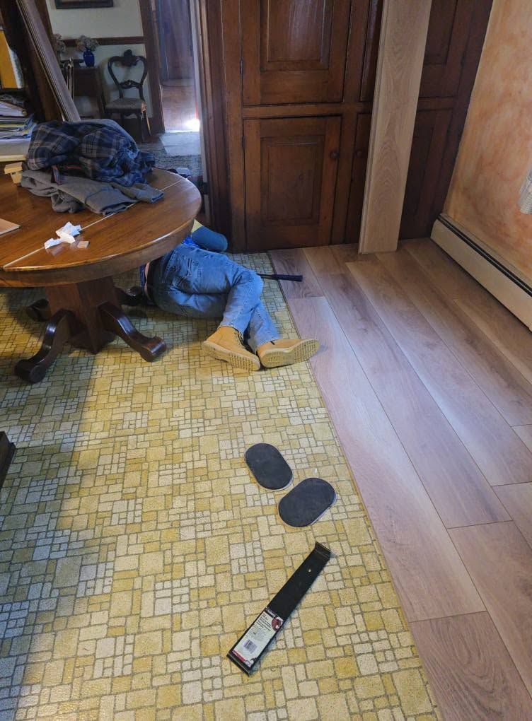 Toddler under a table on a yellow rug, with shoes and a TV remote on the floor nearby.
