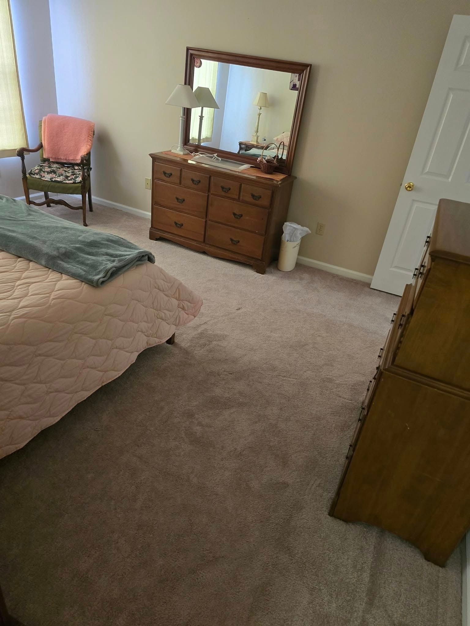 Bedroom with beige carpet, bed, dresser with mirror, chair by window, and white door.