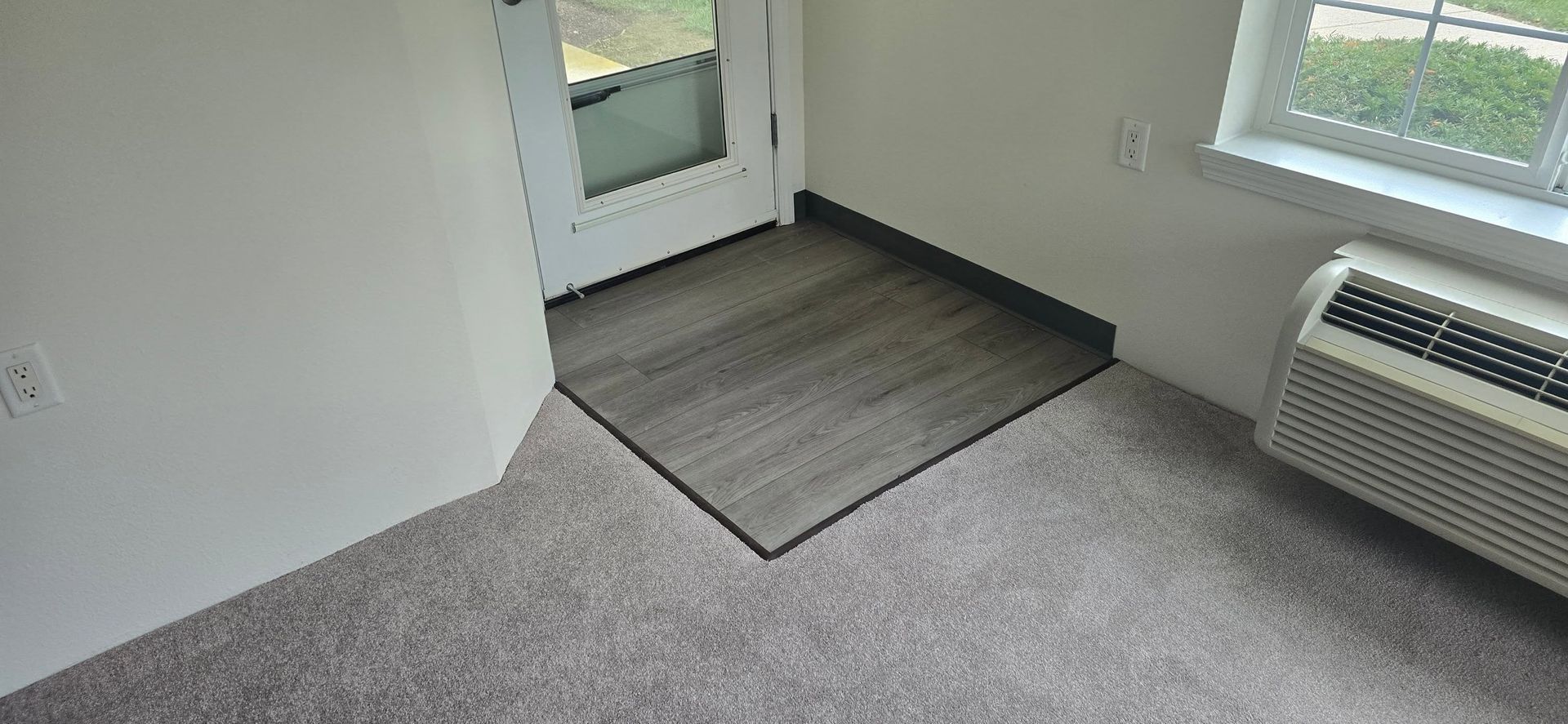 Empty room corner with gray carpet, door mat, window, and wall-mounted heater.