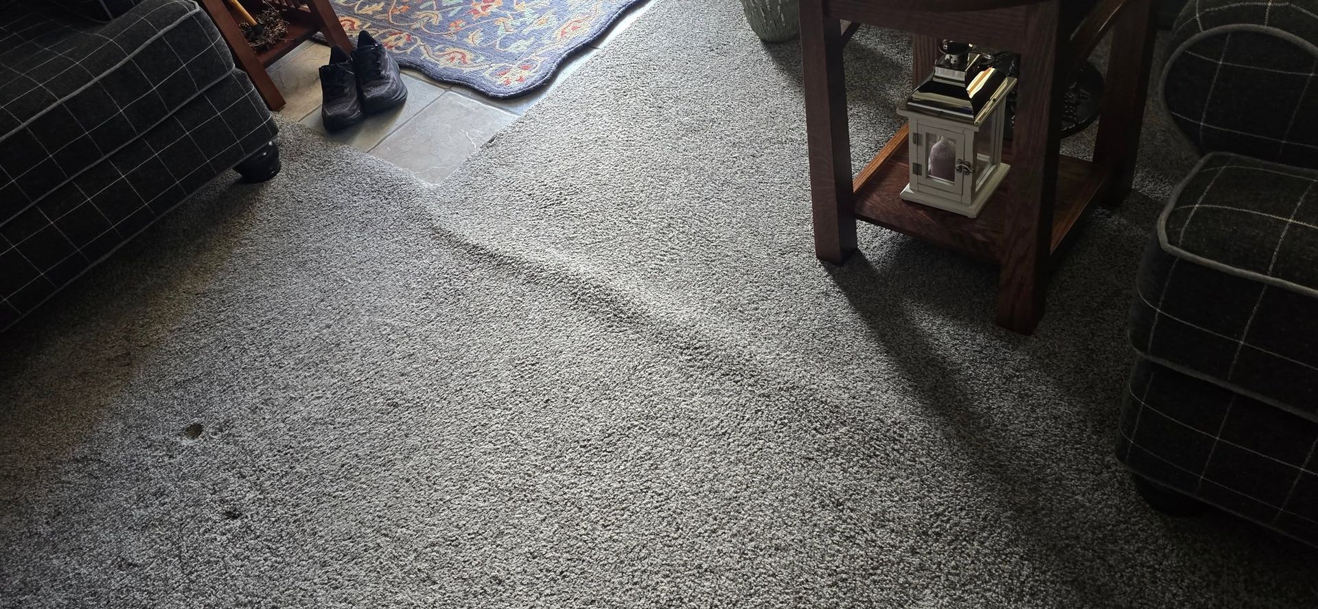 Living room carpet with sunlight, a person’s shoes at top left, and a small wooden side table on the right