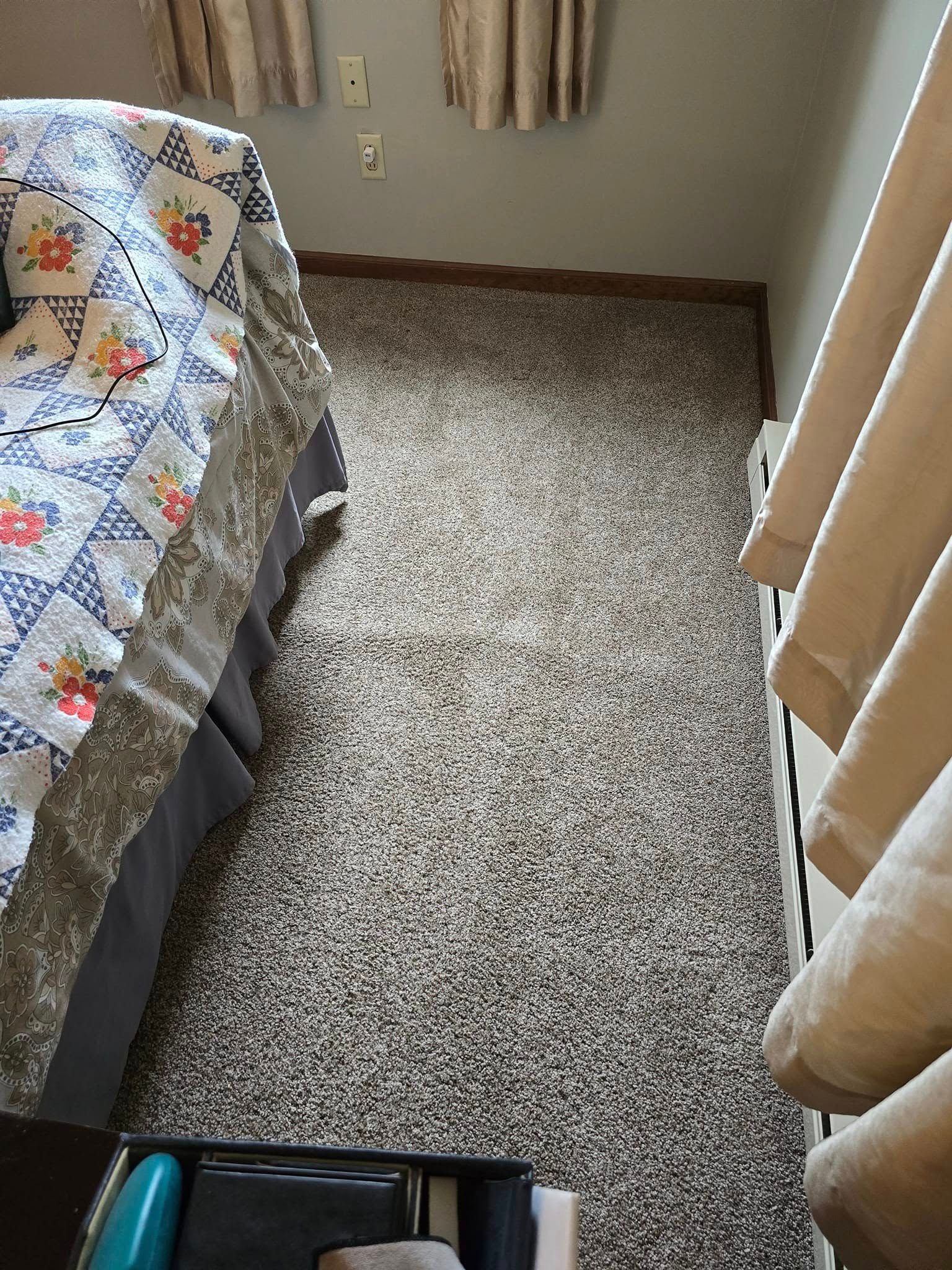 Carpeted bedroom aisle beside a bed with a floral quilt and beige curtains.