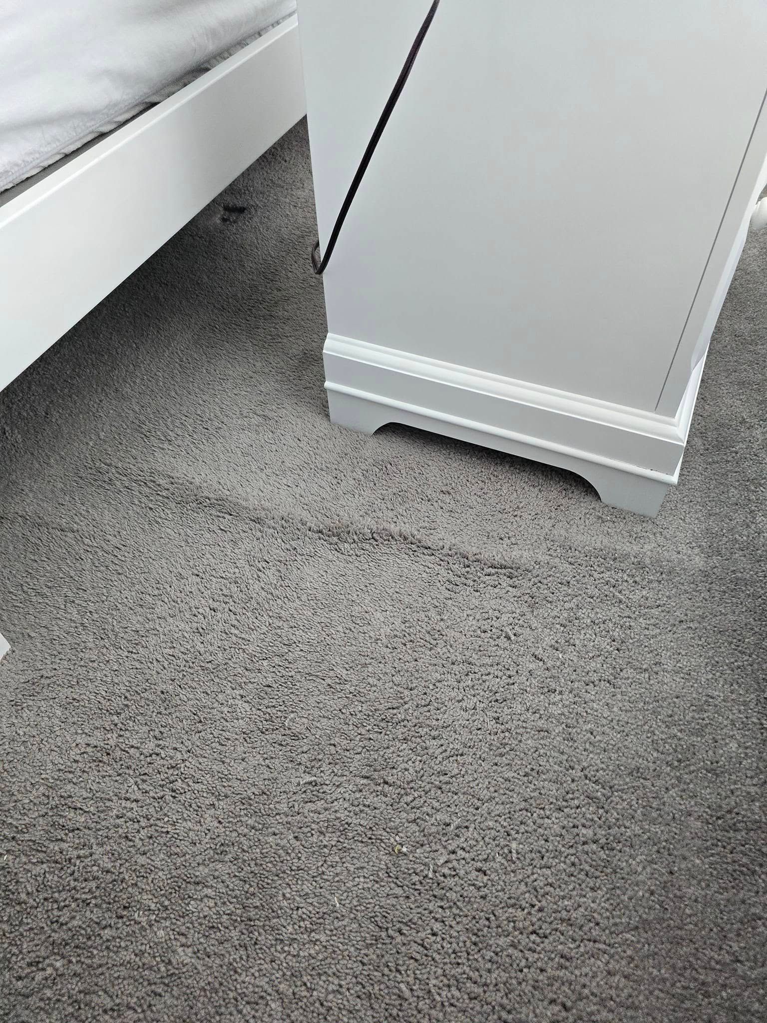 Gray carpet in a room corner beside a white baseboard and wall trim, with a small gap under the trim