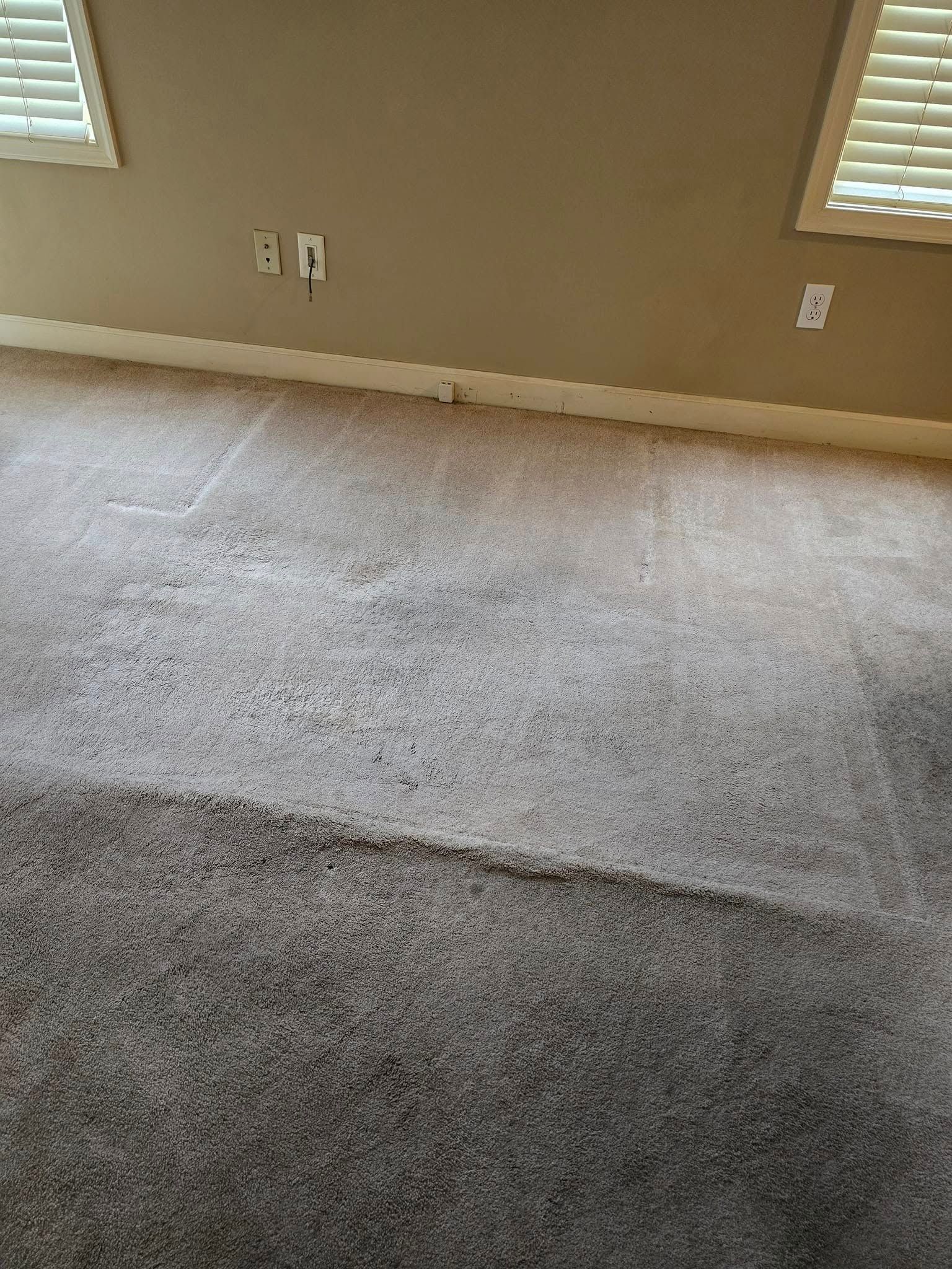 Empty beige-carpeted room with tan walls, two windows, and electrical outlets along the baseboard.