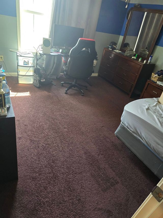 Cluttered bedroom with red carpet, desk chair, dresser with mirror, and bed by the window.