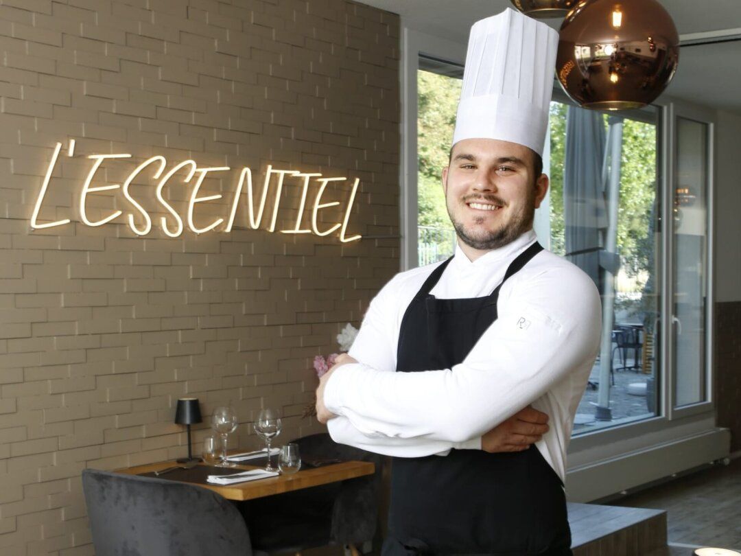 Chef en uniforme blanc et tablier noir, debout près d’une enseigne au néon « L’ESSENTIEL » dans un restaurant.