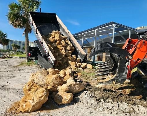 Dump truck unloading rocks near a pool enclosure, with an excavator present on a sunny day.