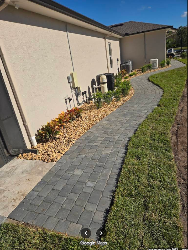 Brick walkway curving along a beige house with landscaping and green grass.