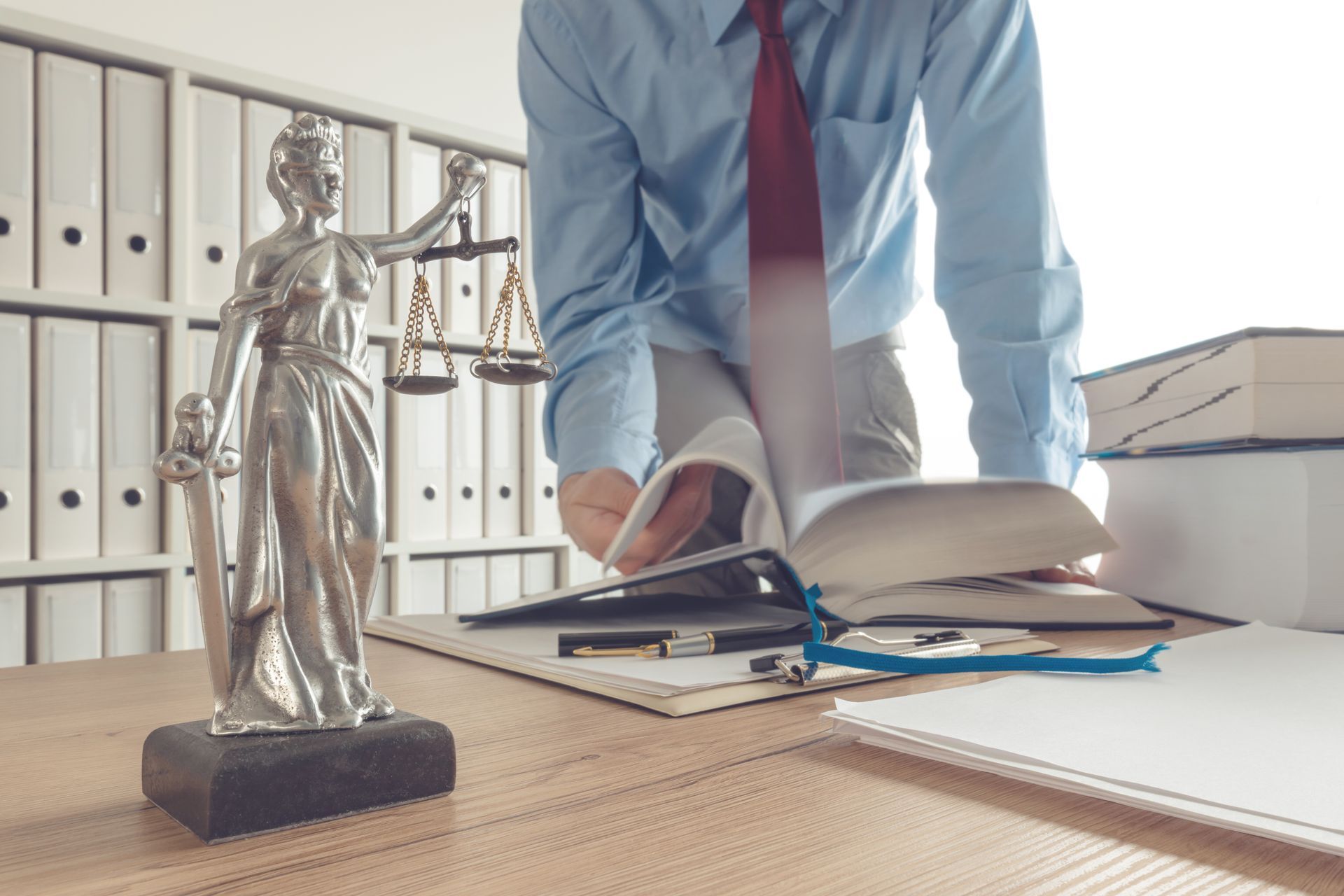 A lawyer in a blue shirt and burgundy reads law books with the justice statue beside them,