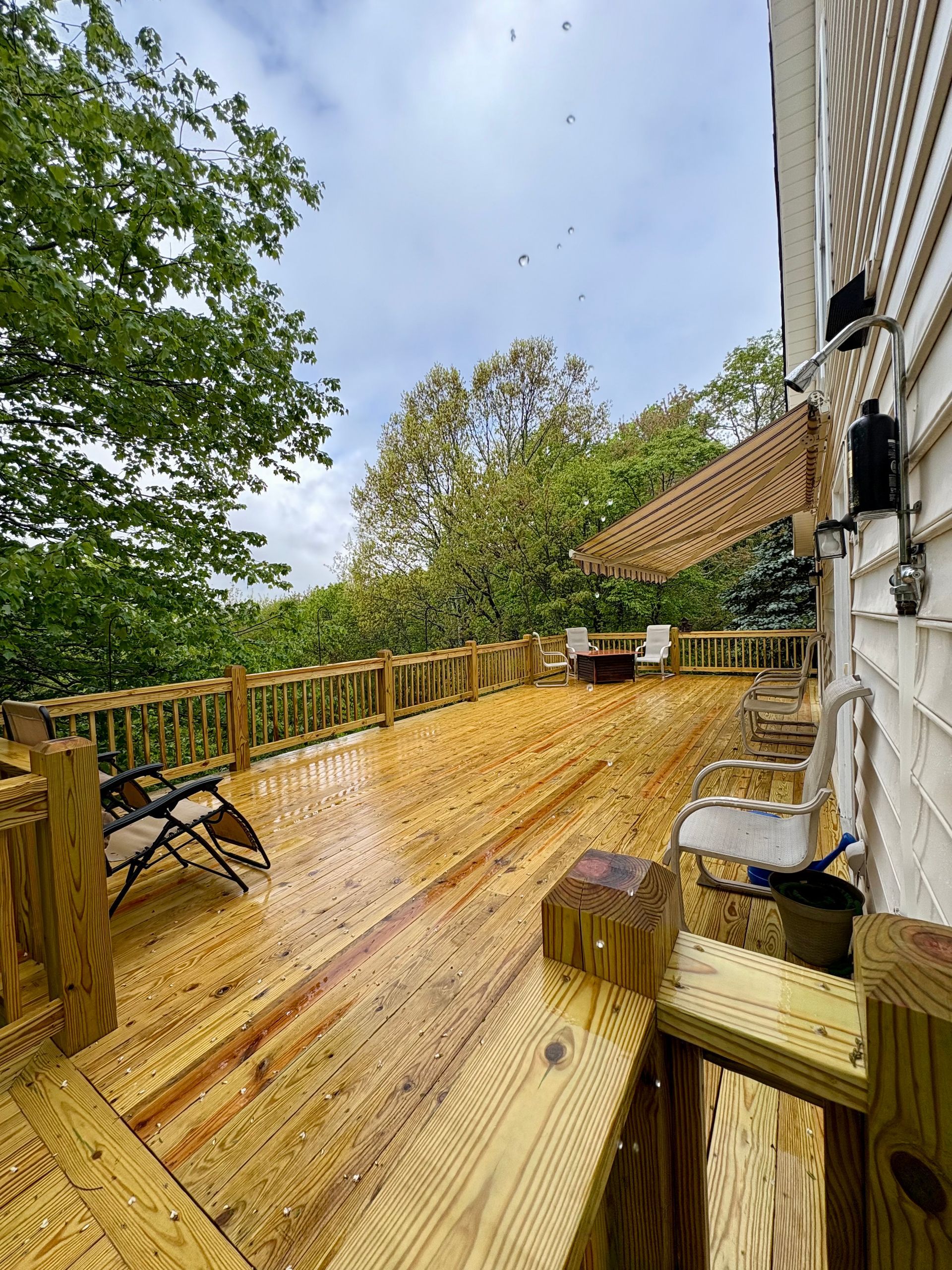 Wooden deck with railing, awning, and outdoor furniture, next to a white house under a cloudy sky.