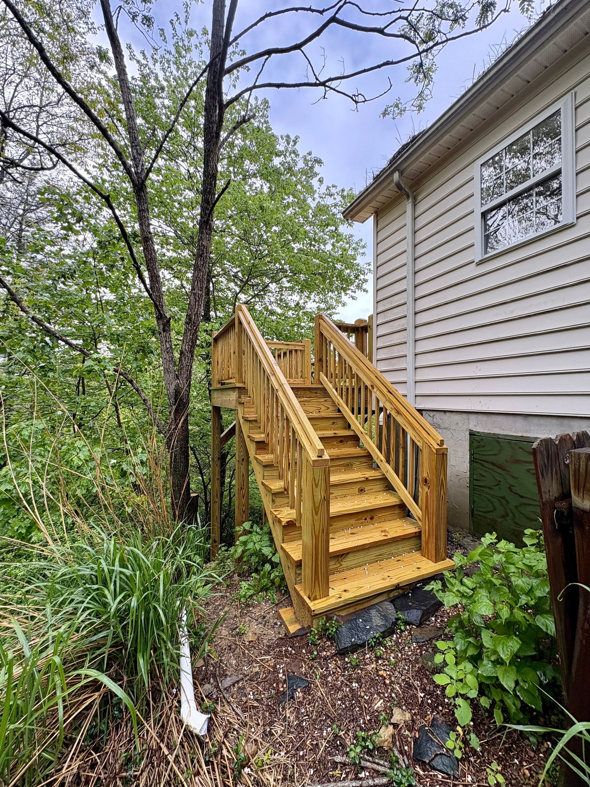 Wooden outdoor staircase leading up to a light-colored house with a window, surrounded by trees and foliage.