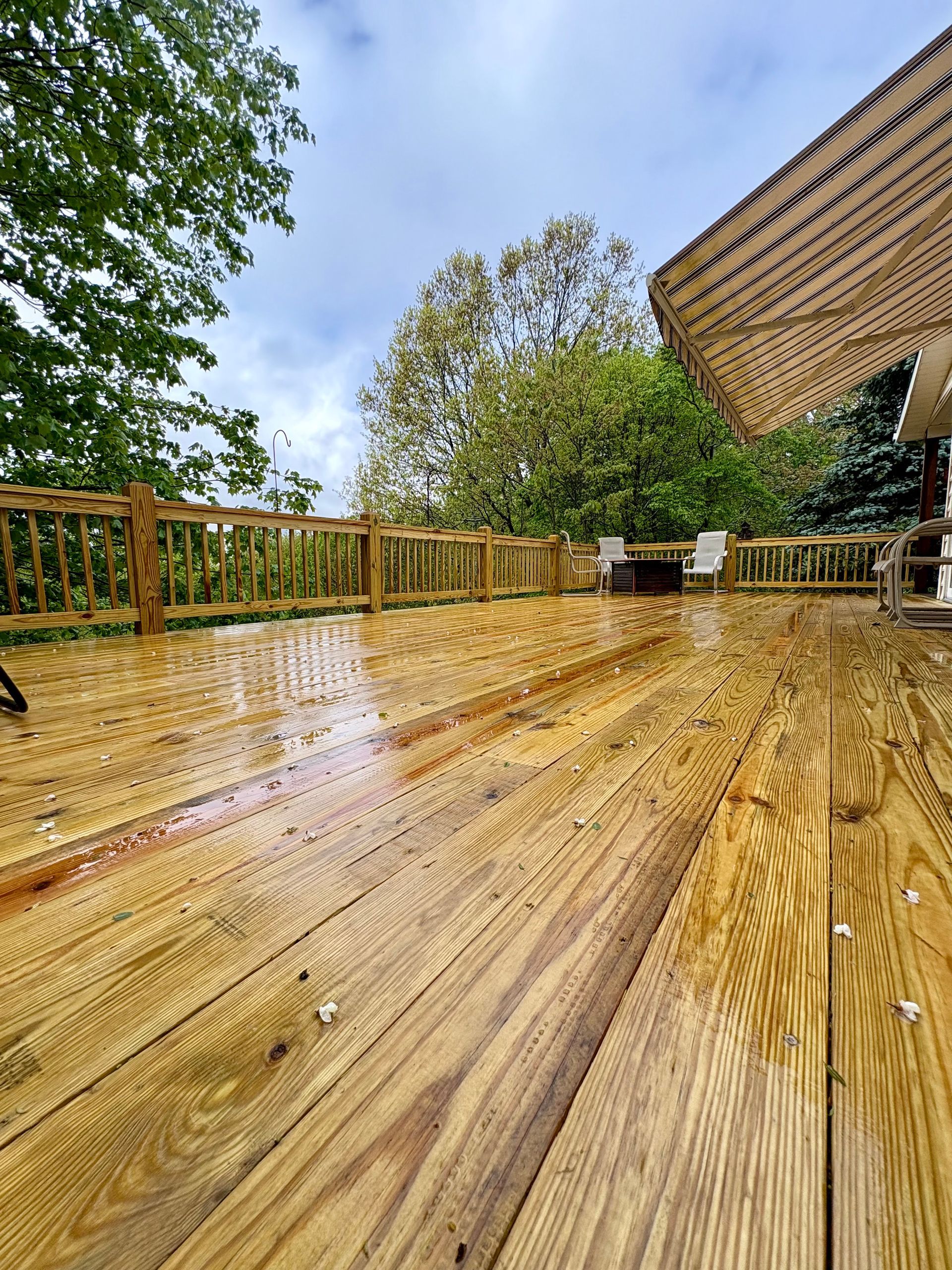 Wet wooden deck with a railing and partial awning, trees in background, overcast sky.