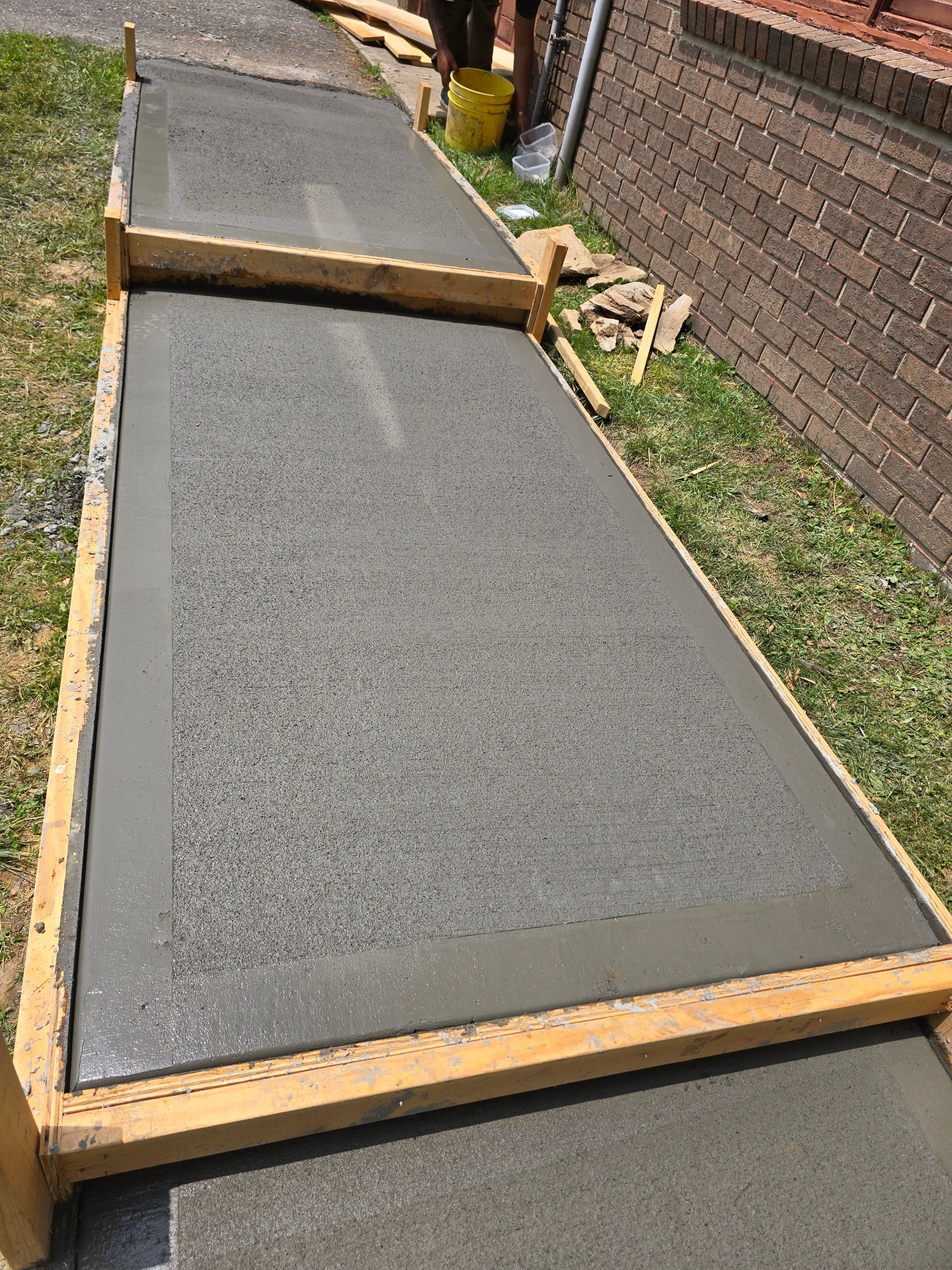 Freshly poured concrete sidewalk within wooden forms, beside a brick wall and grass.
