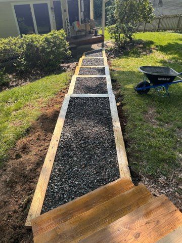 Wooden steps filled with black gravel, built in a grassy yard, leading to a porch. A wheelbarrow sits nearby.