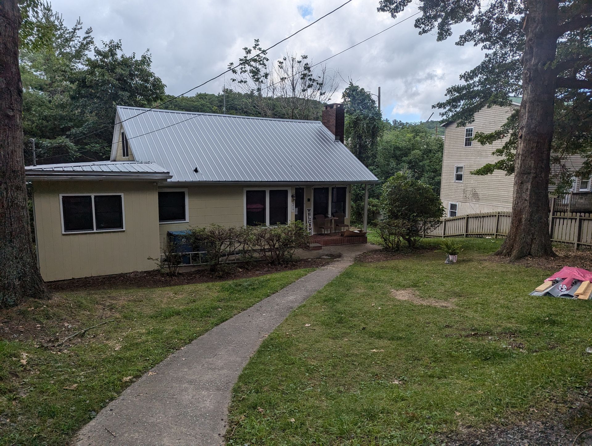 Small beige house with a metal roof, concrete path, and yard. Trees frame the house on a cloudy day.