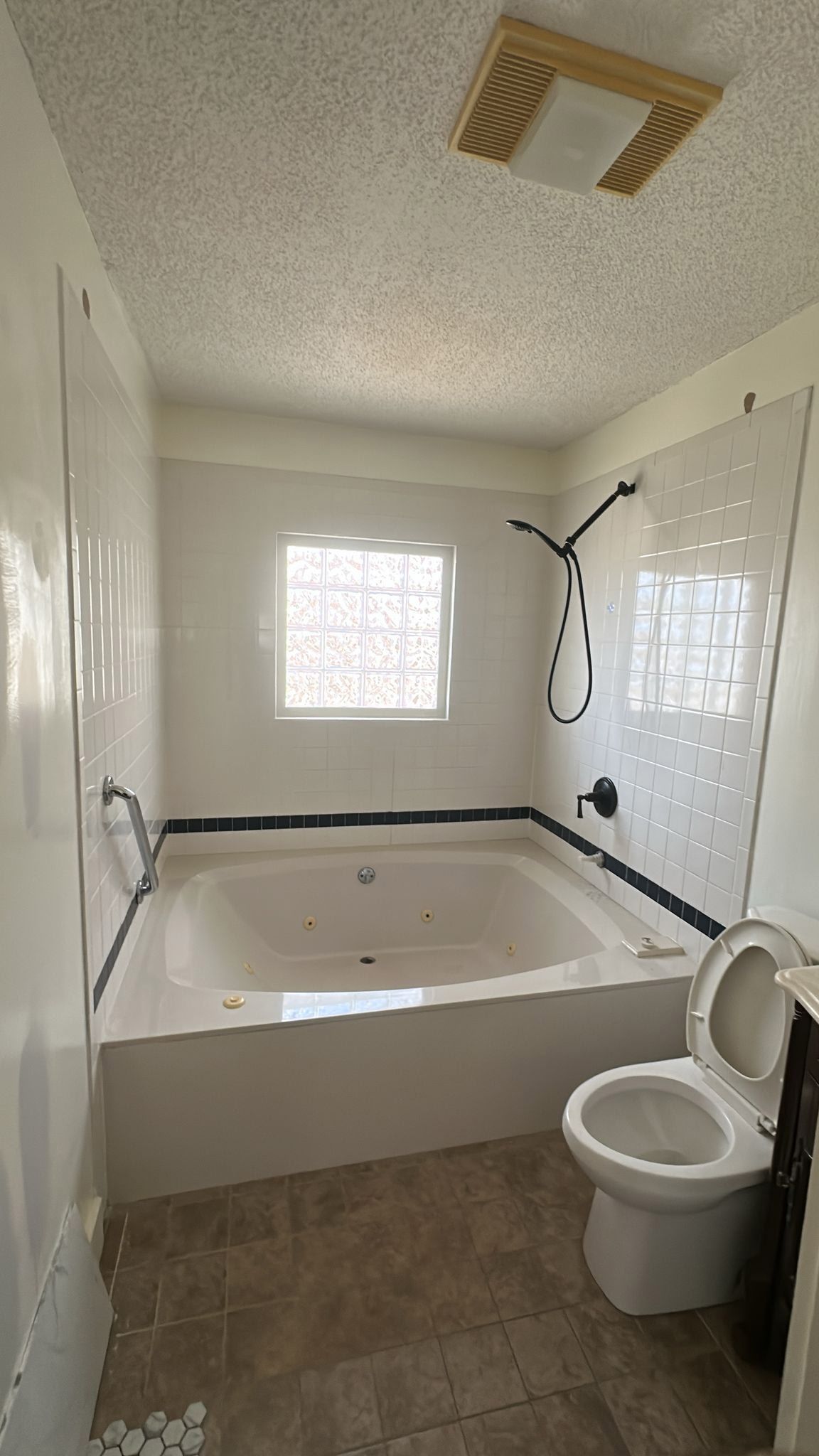 Bathroom with white walls, tiled tub surround, black shower fixtures, toilet, and window with patterned glass.