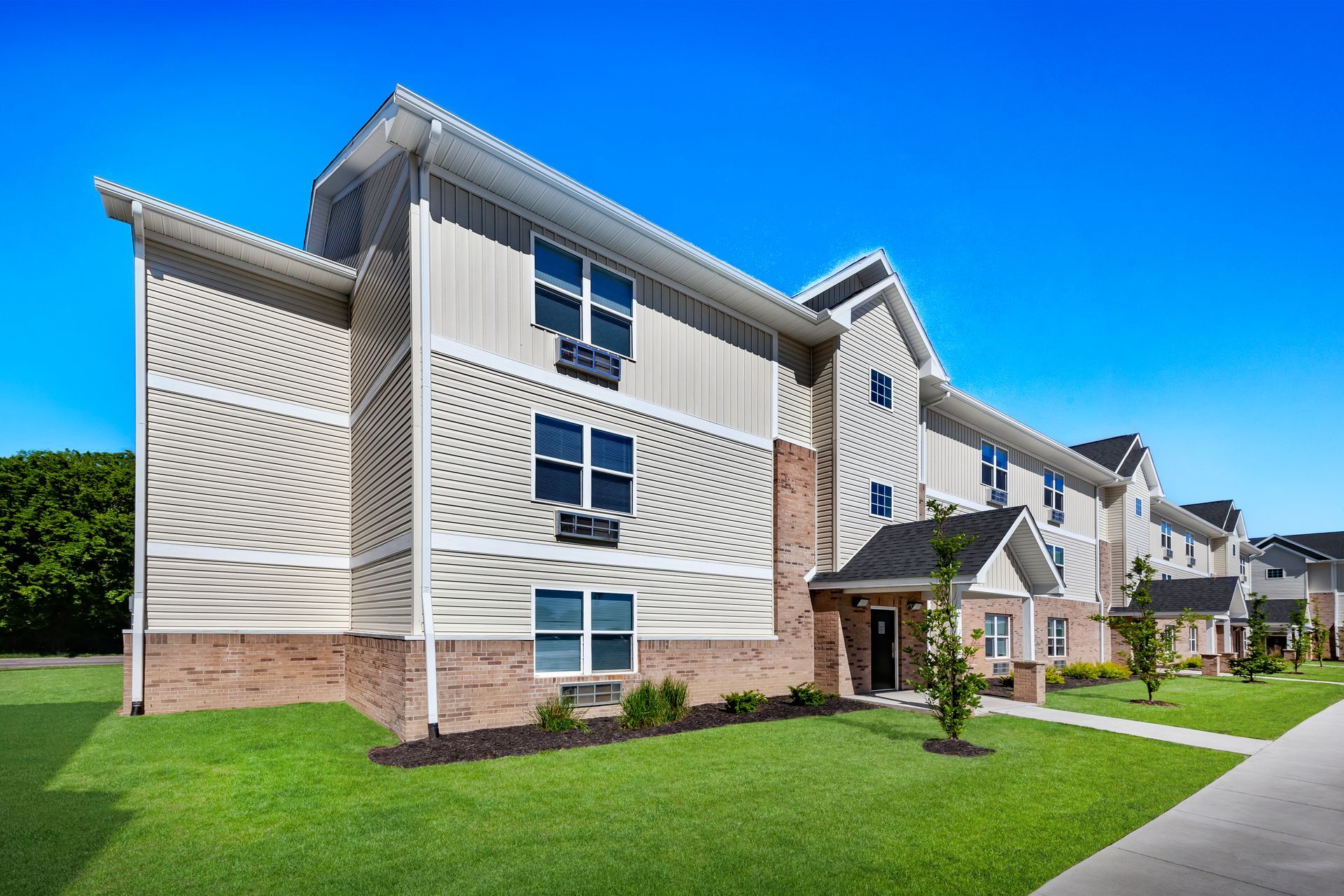 Modern apartment buildings with white siding and brick accents beside a green lawn and sidewalk.