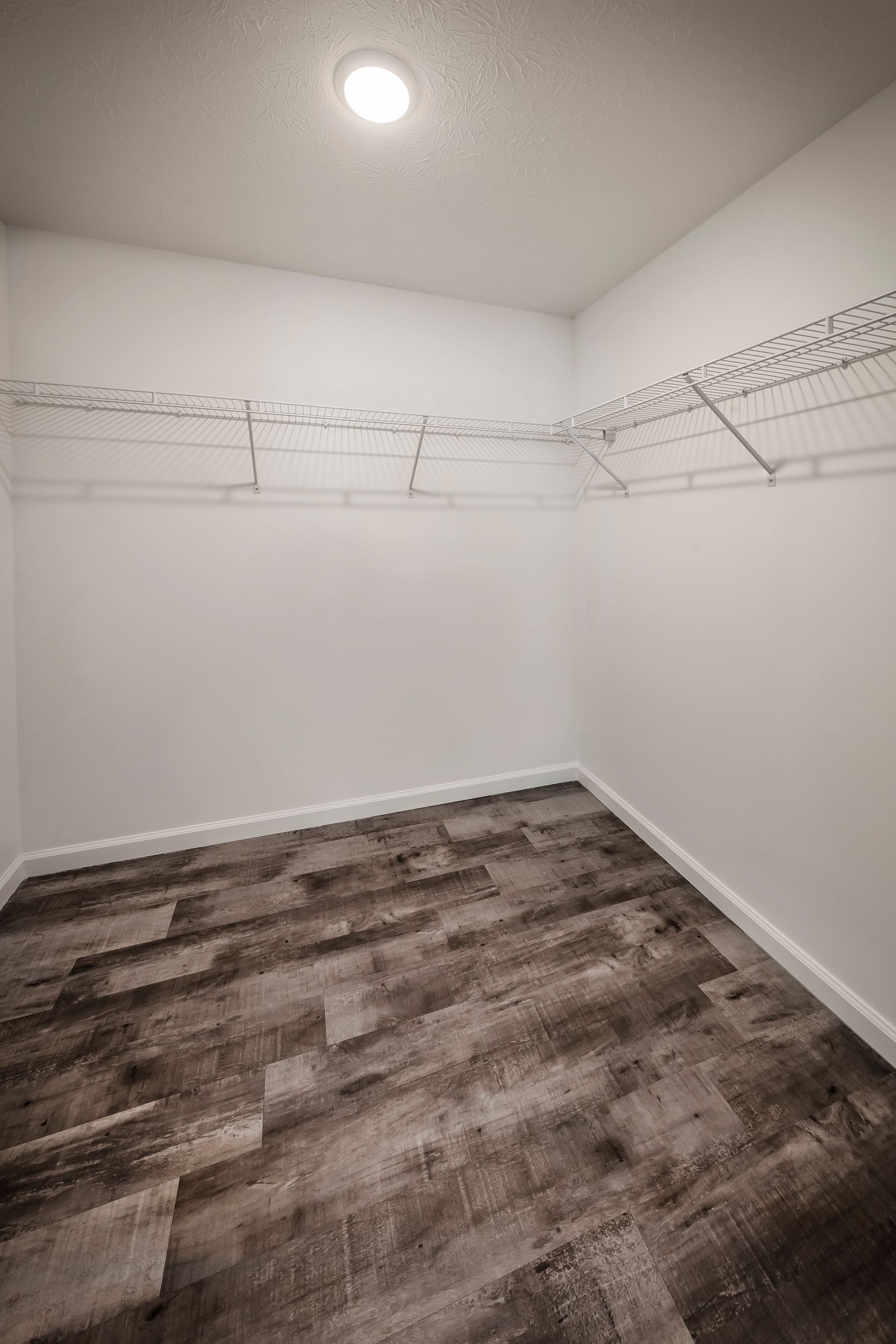 Spacious walk-in closet with white walls, wire shelving, and wood-look flooring under a ceiling light