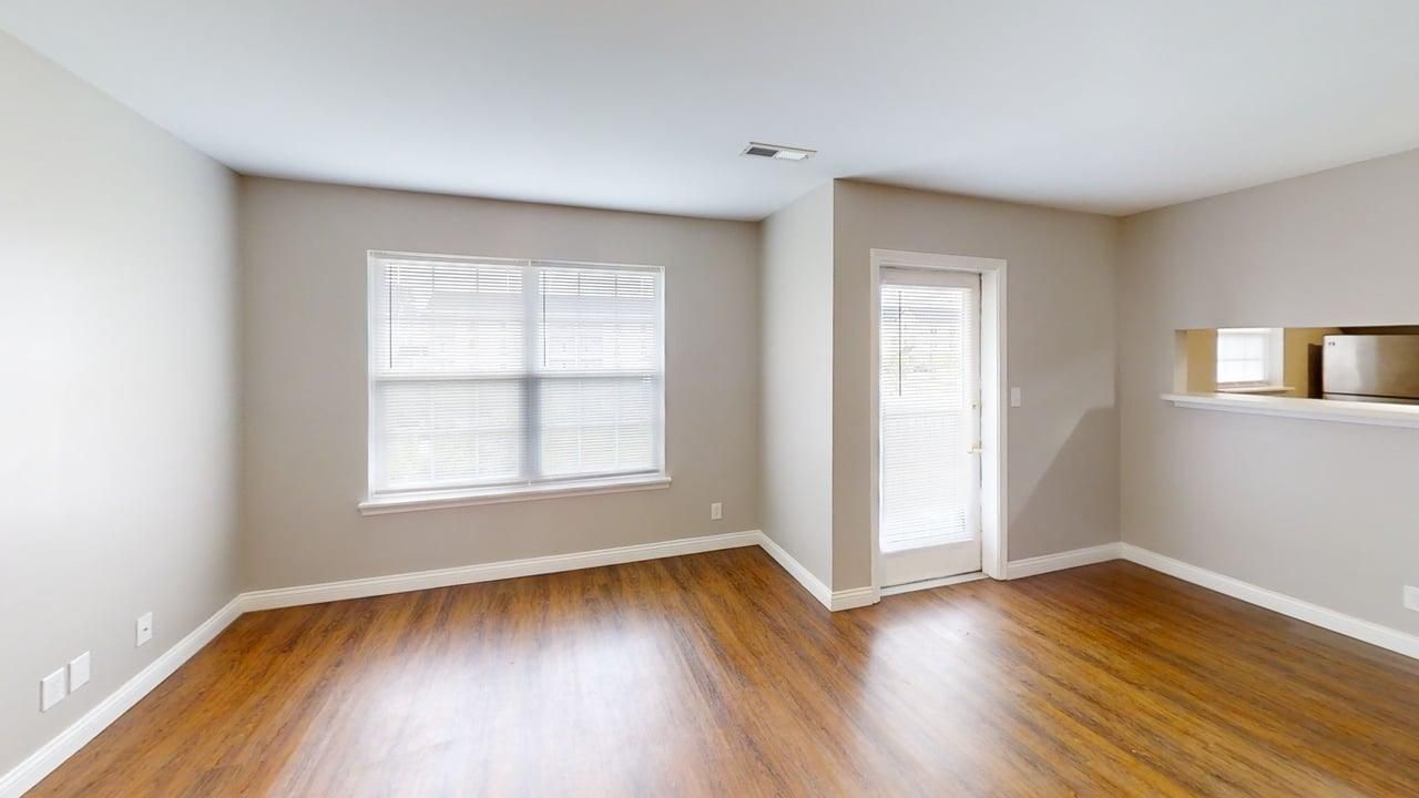 Empty beige living room with wood floors, two windows, and a white door leading outside