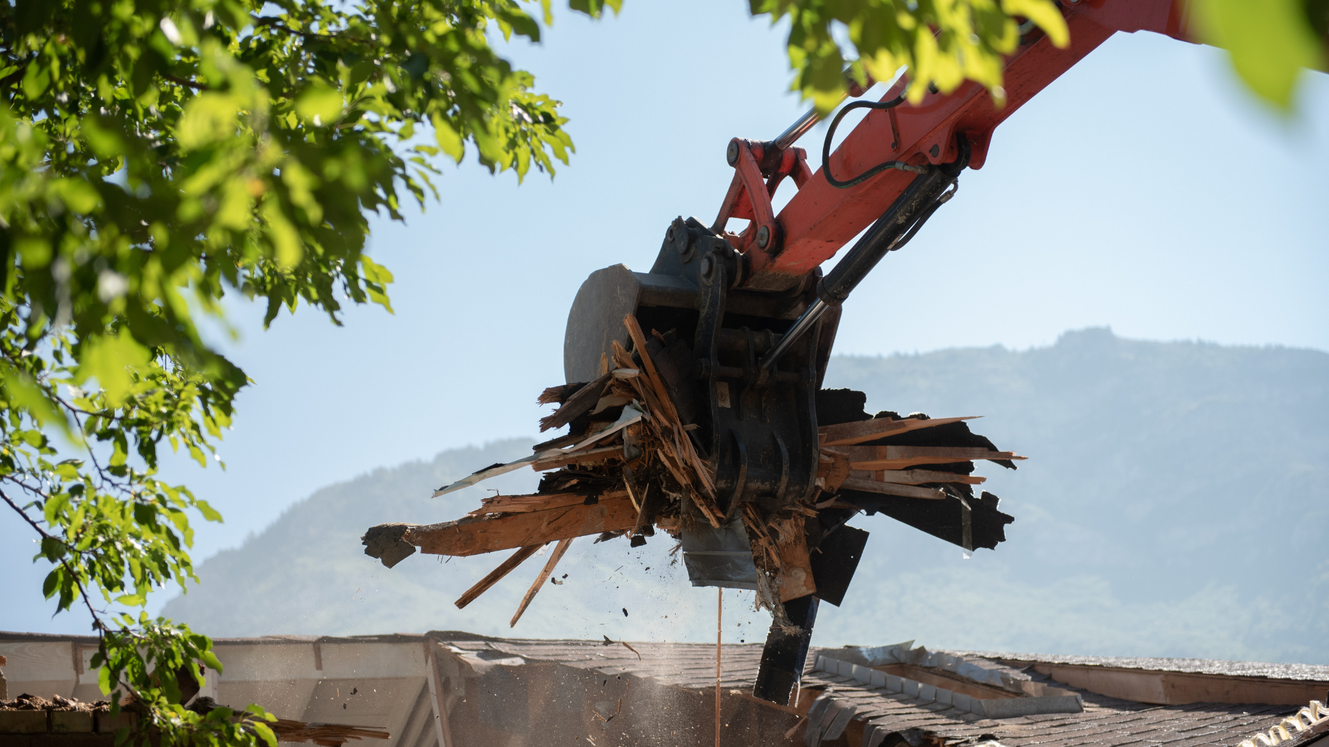 Excavator demolishing a wooden structure, with mountains in the background and leaves framing the top left.