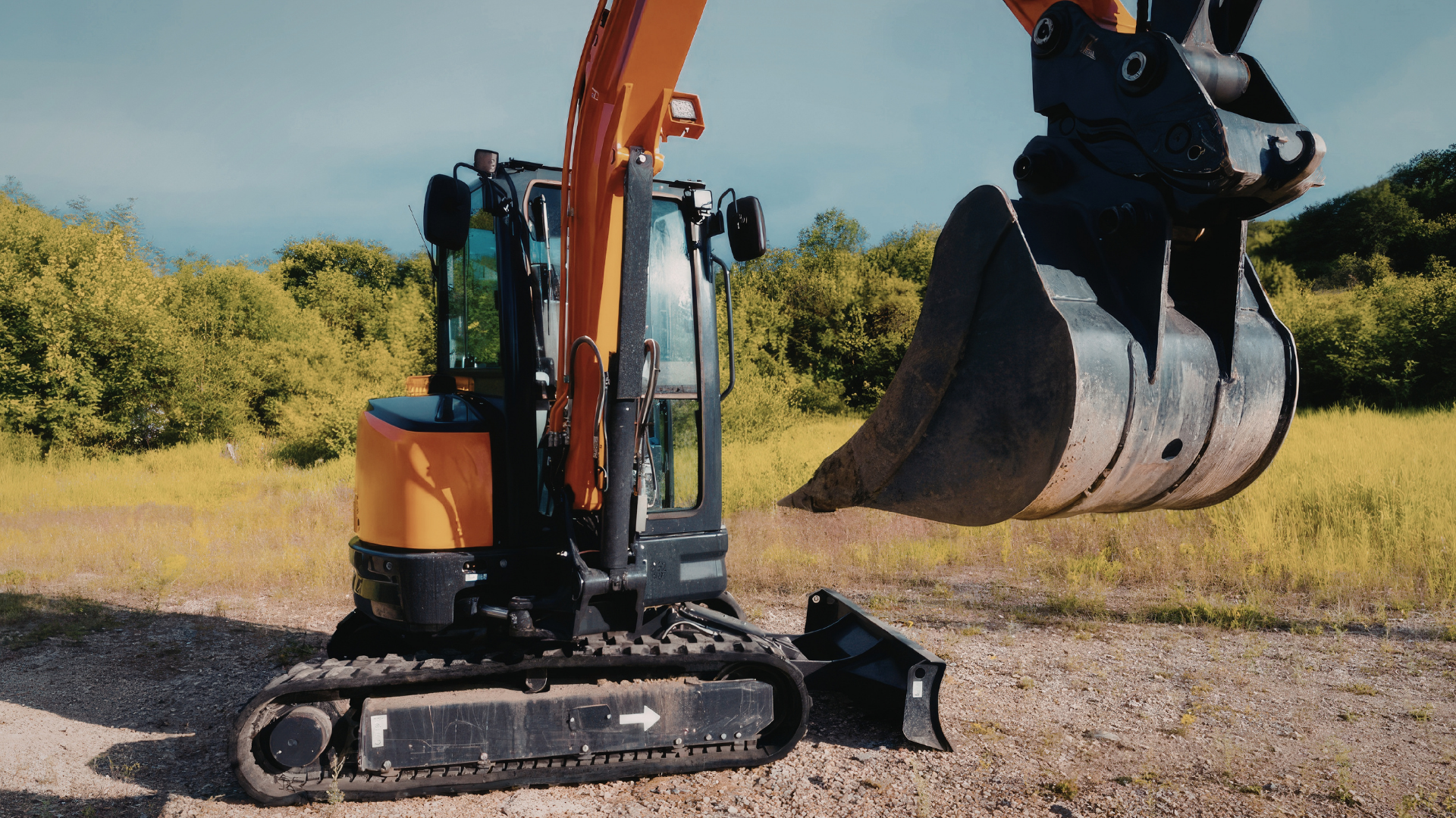 Orange excavator on tracks with raised bucket in a grassy field under a blue sky