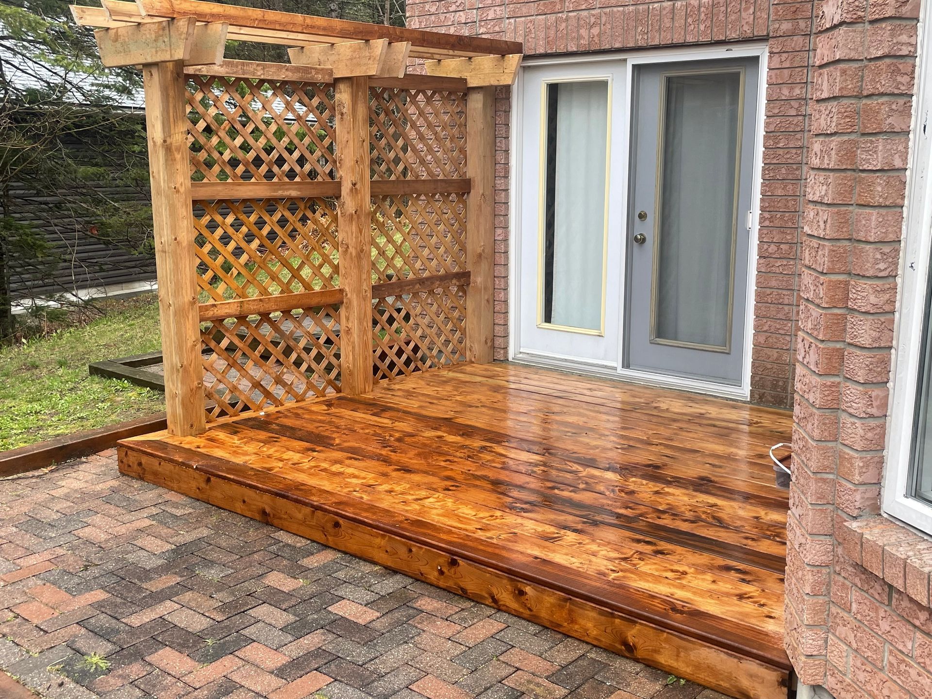 A wooden deck with a pergola and a brick building in the background.