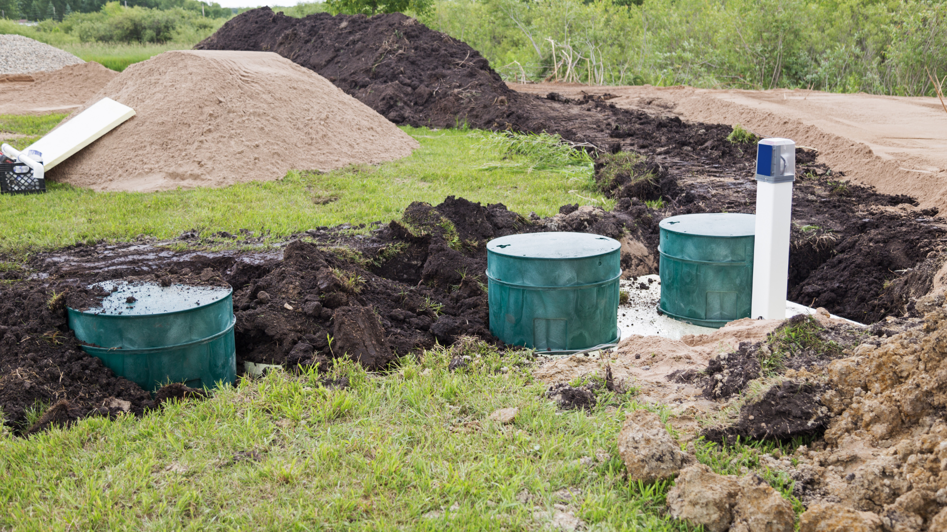 Three green septic tanks installed in a grassy area with dirt and sand piles.
