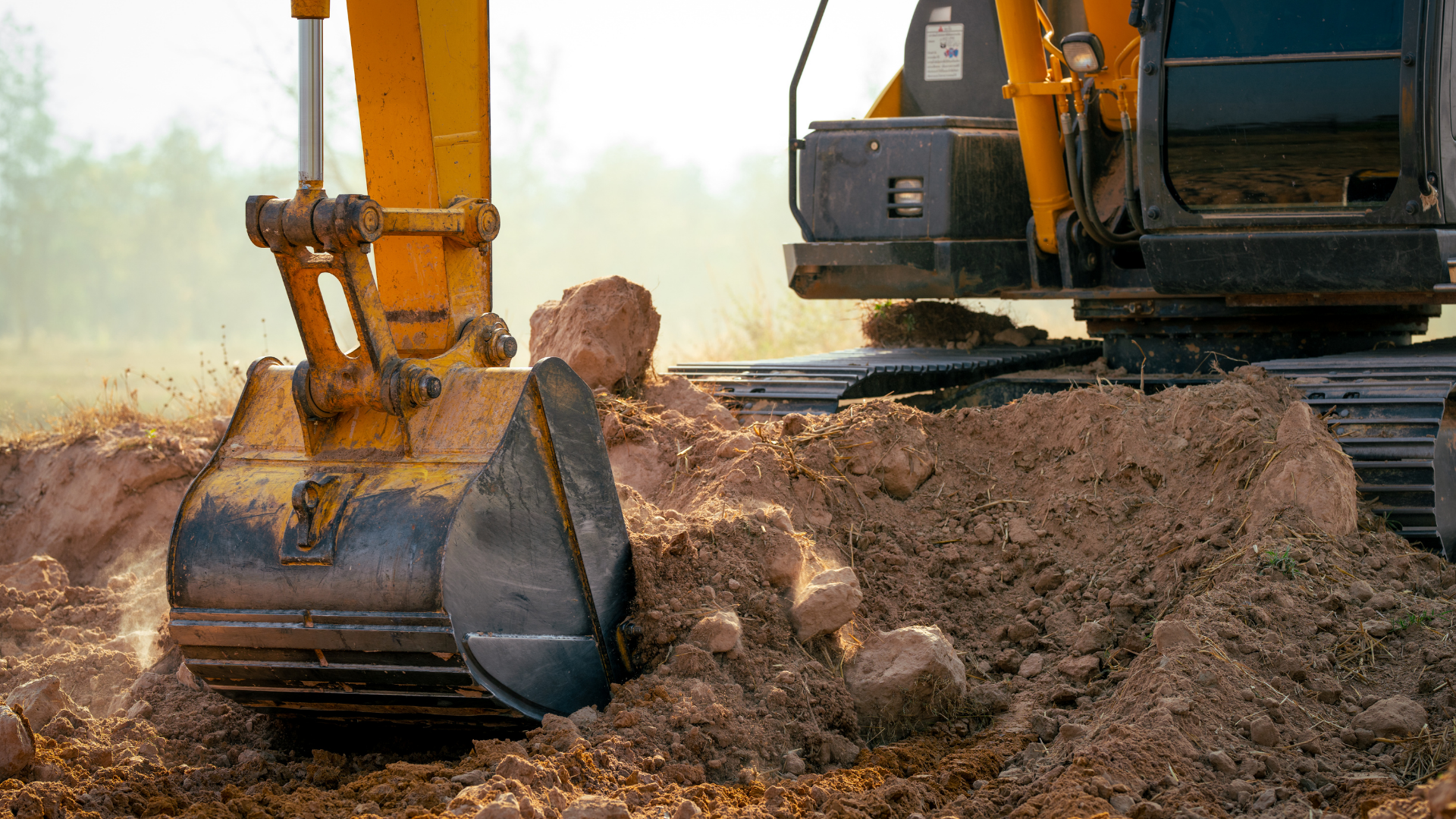 Excavator bucket digging soil at a construction site with a dirt mound and machine tracks.