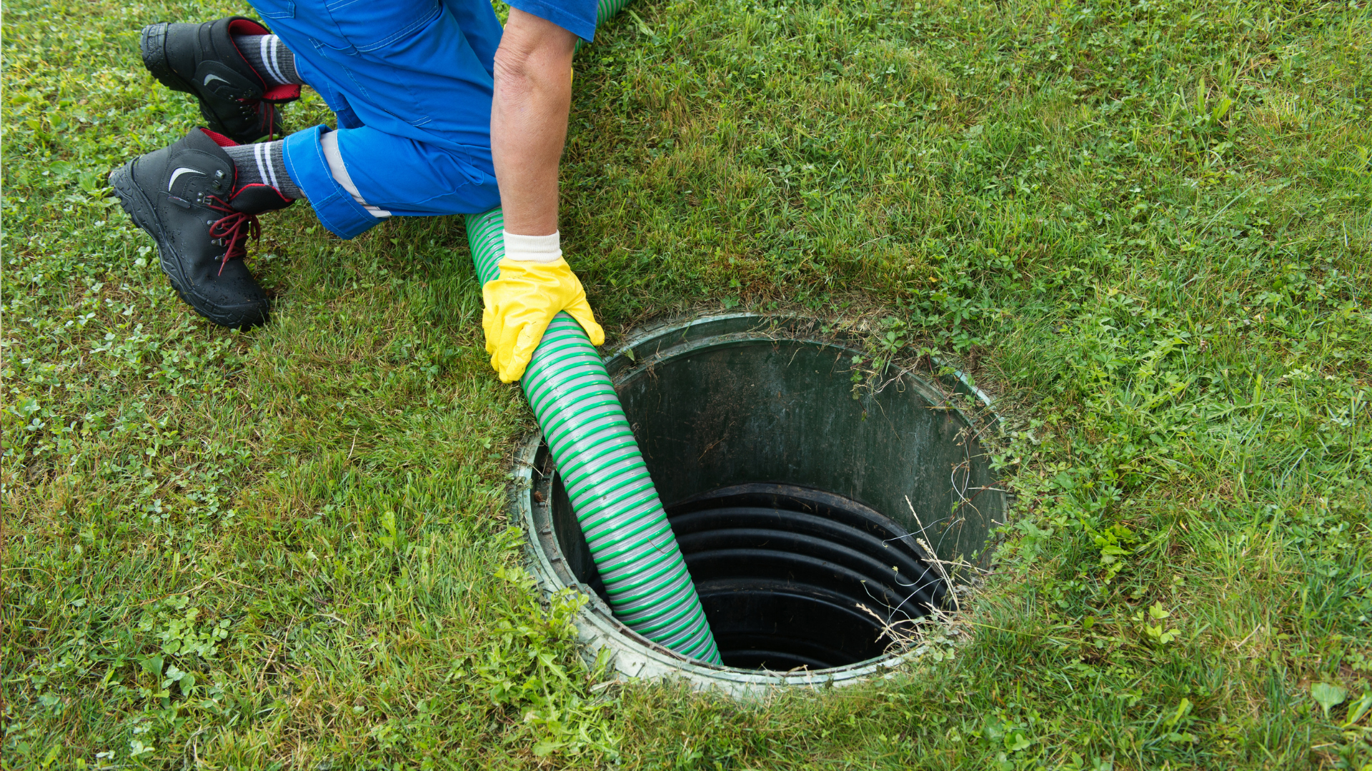 Worker in blue pants using a green hose to clean a drain opening in the grass