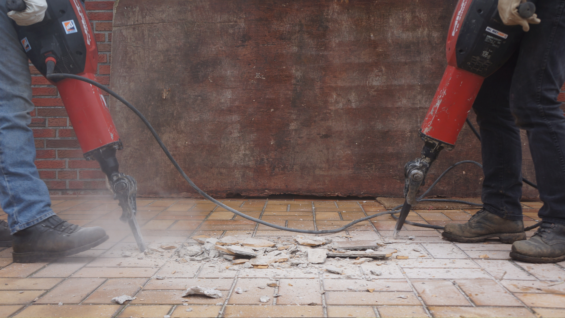 Two workers using red jackhammers to break concrete floor near a wall, with dust and debris rising.