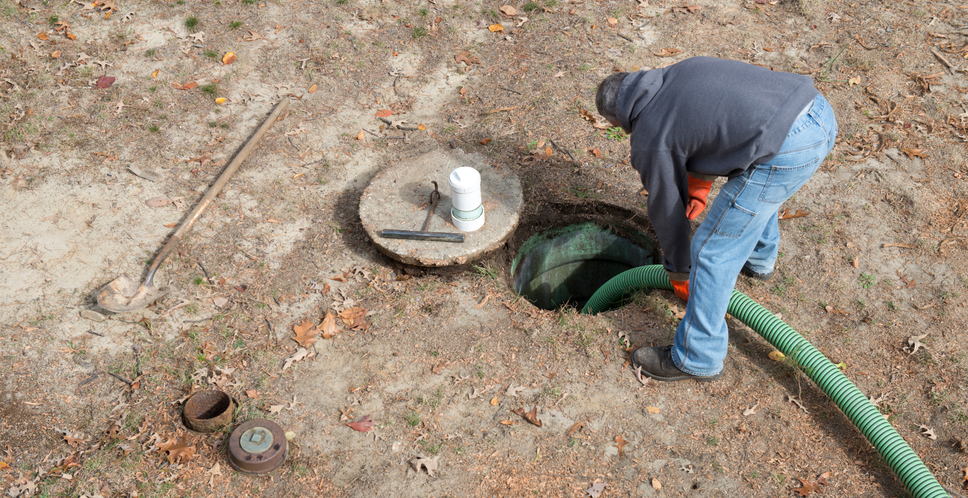 Worker inspecting an open utility hole with a green hose in a dirt yard
