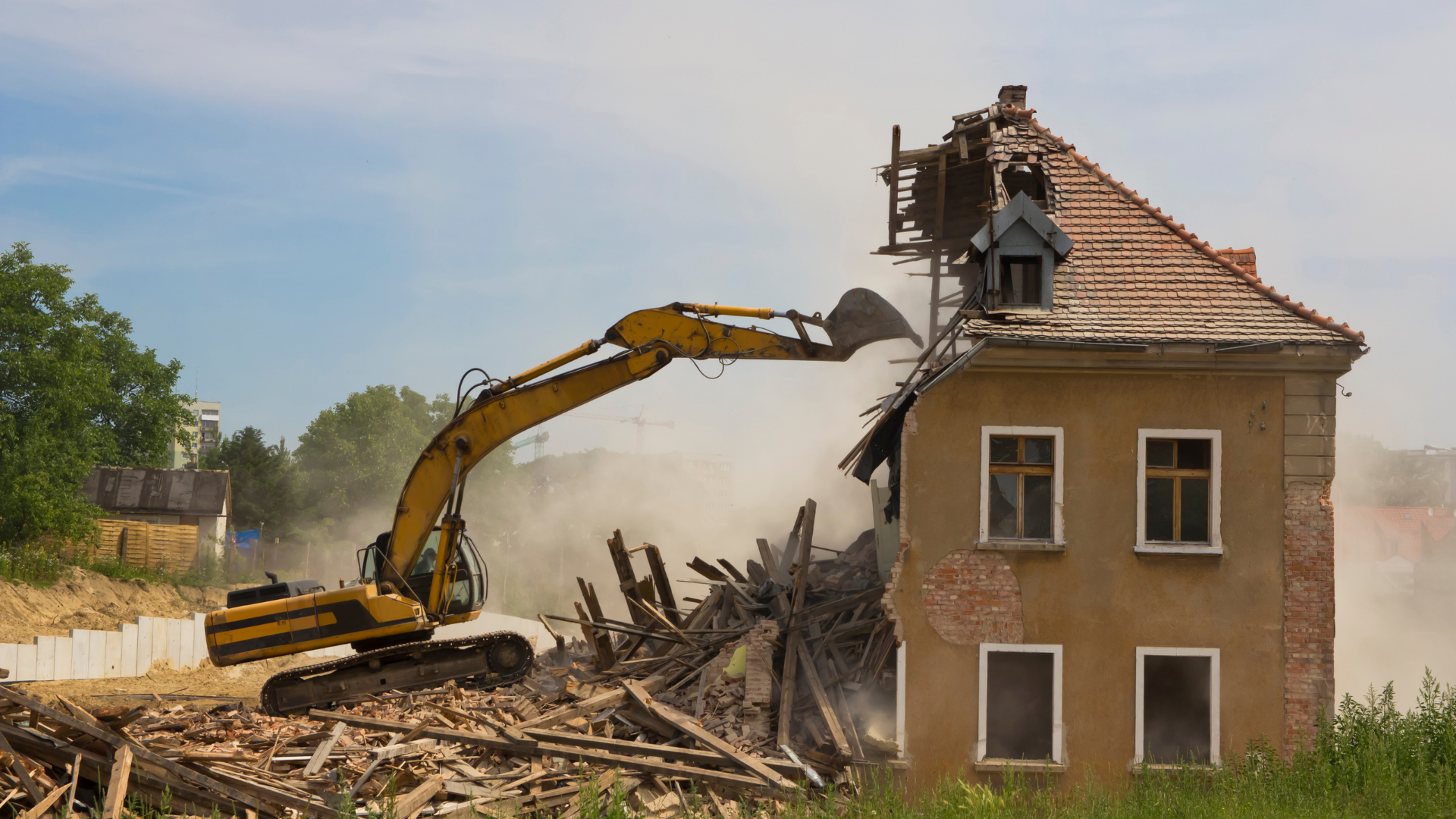 A yellow excavator tears down the side of an old, multi-story building, creating a cloud of dust and debris.