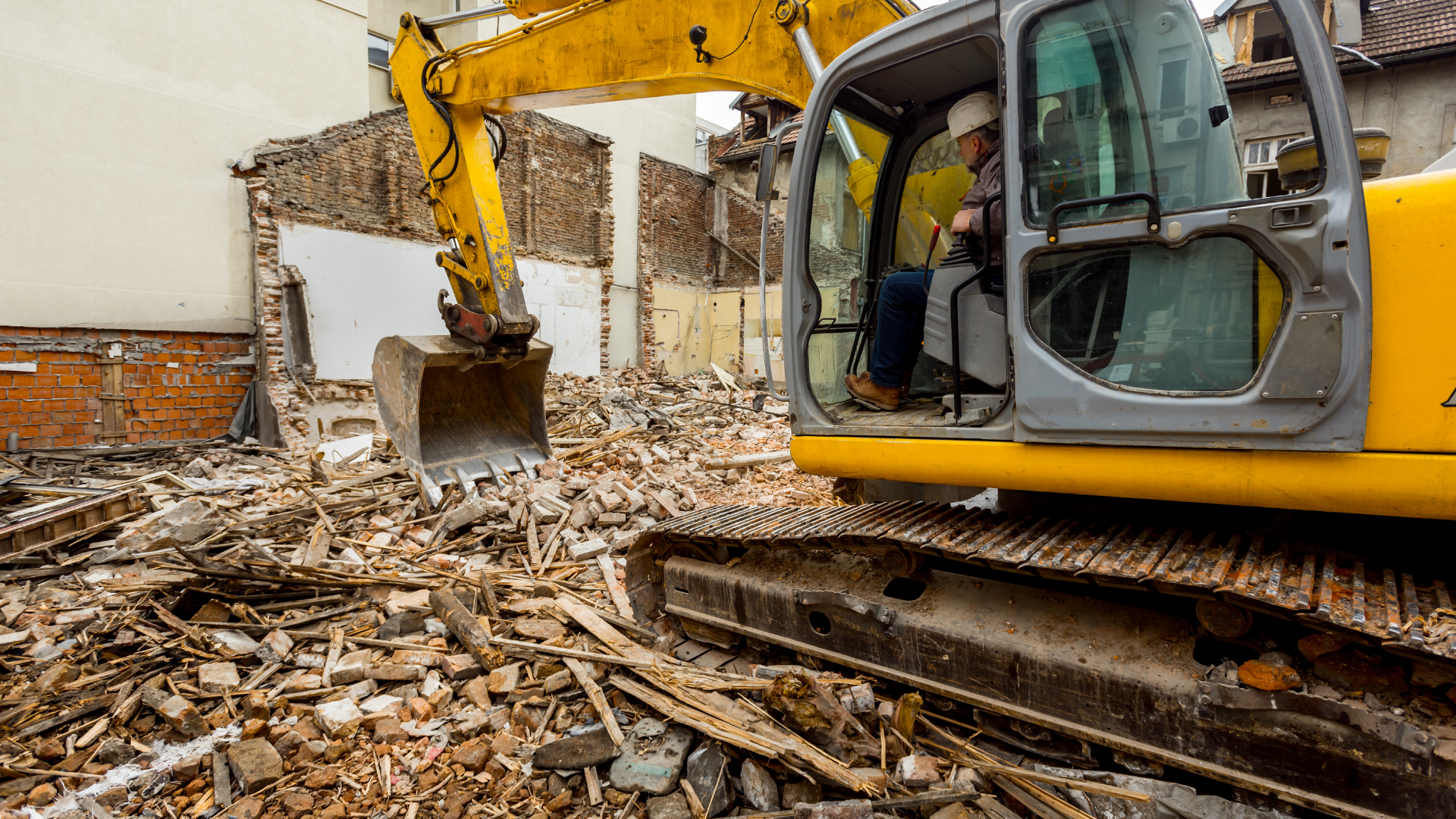 Yellow excavator demolishing a building; operator in cab. Debris surrounds the machine.