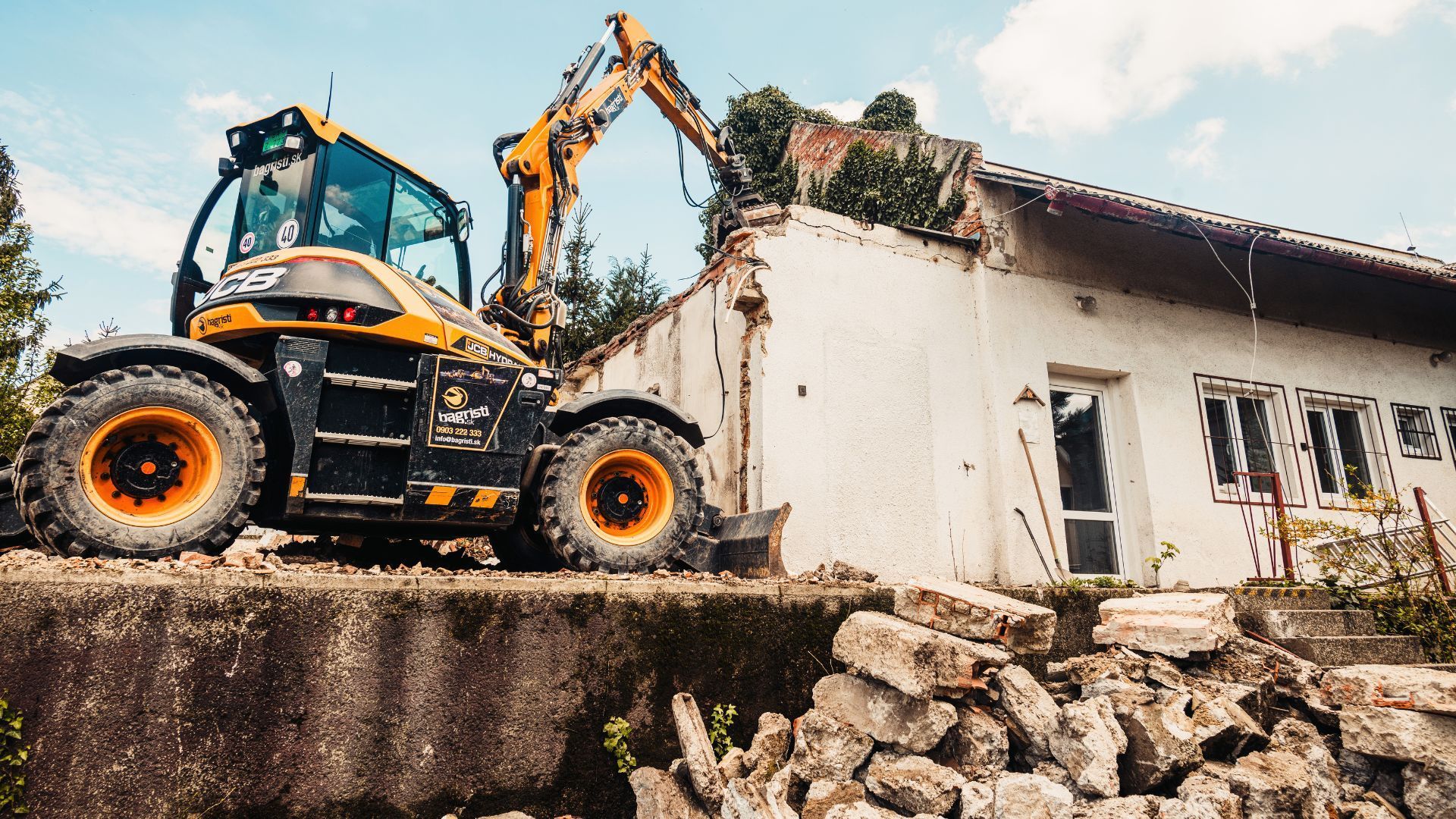 Excavator demolishing a white house beside a rubble pile on a construction site