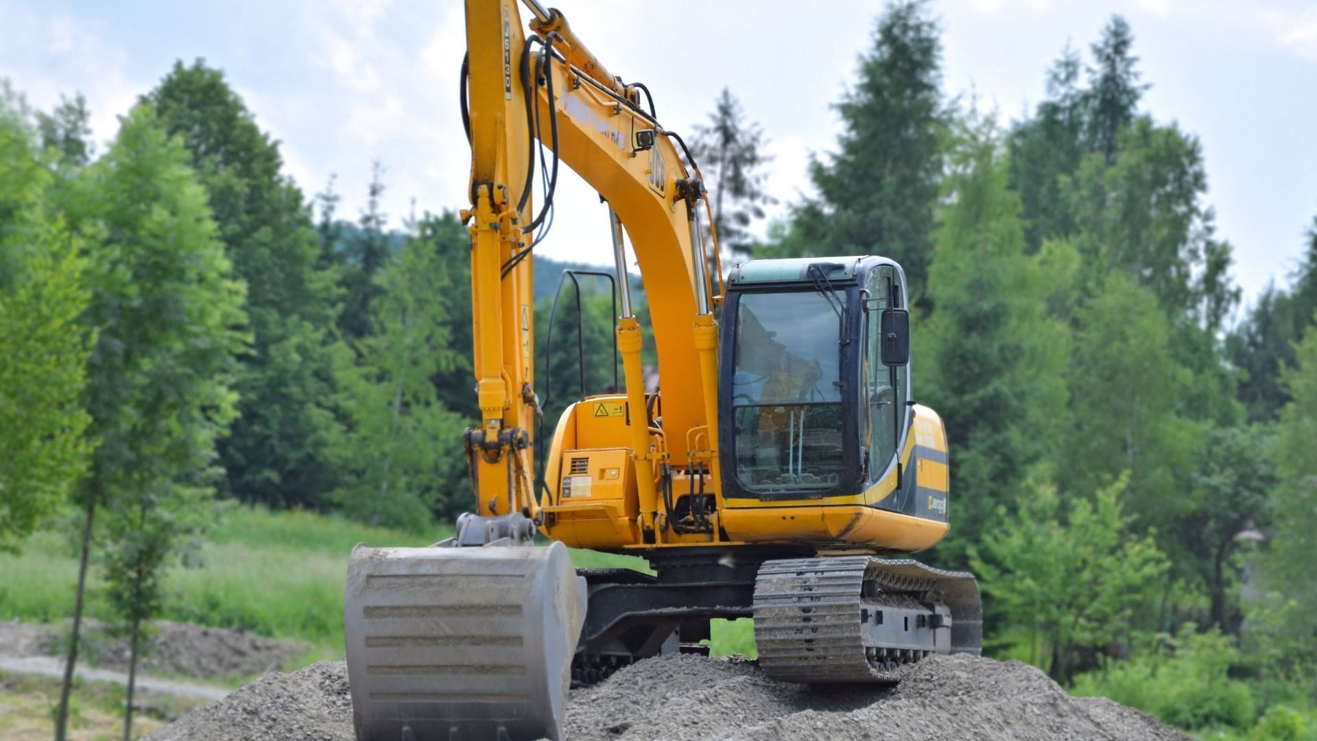 Yellow excavator parked on a gravel pile beside a forested path.