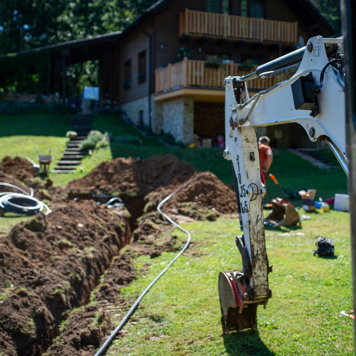 A white excavator stands in a grassy yard beside a long trench being dug in front of a rustic wooden house.